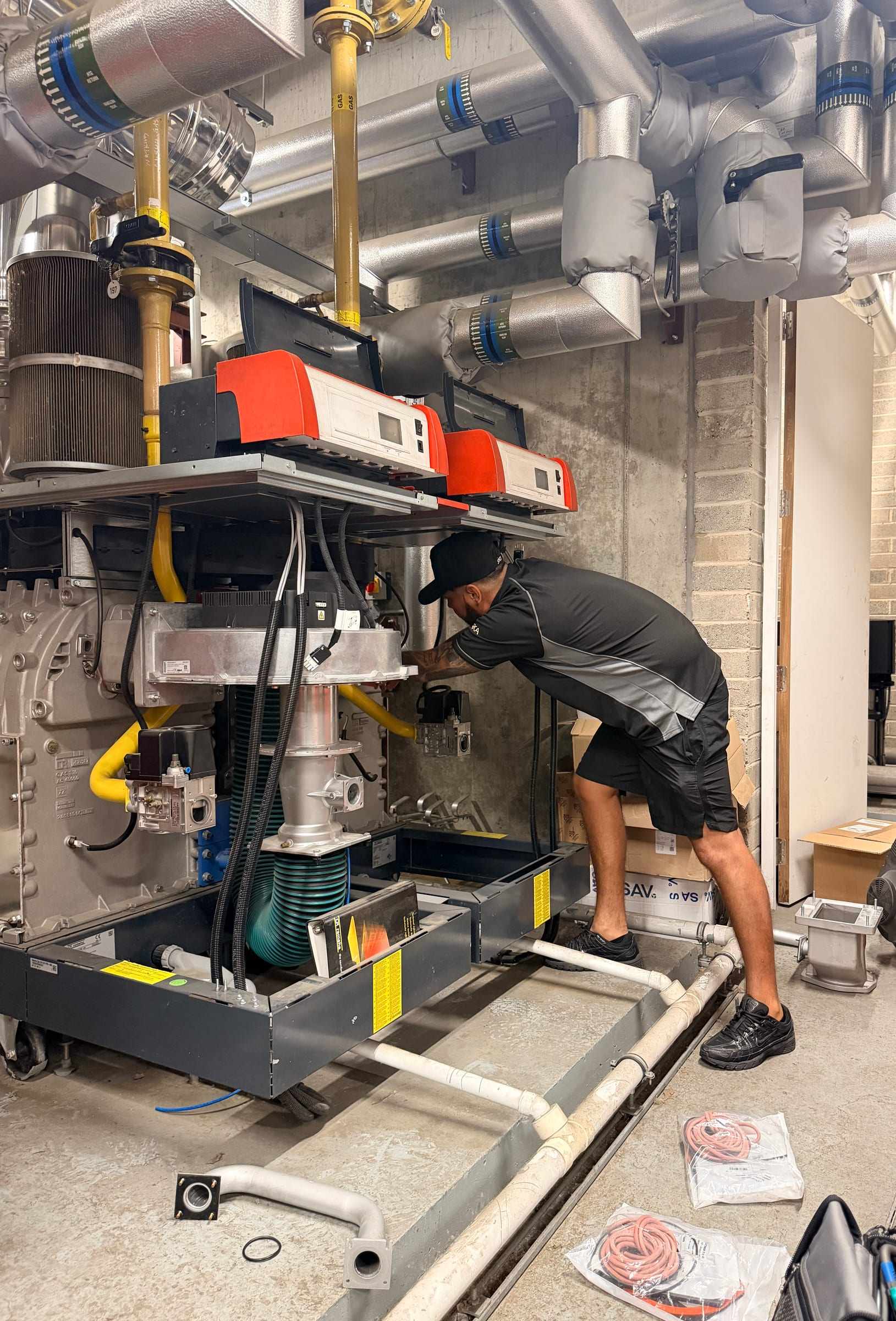 A Neos Engineering engineer working on the gas controls of commercial boiler plant in a London plant room, under insulated heating pipes and yellow gas pipework
