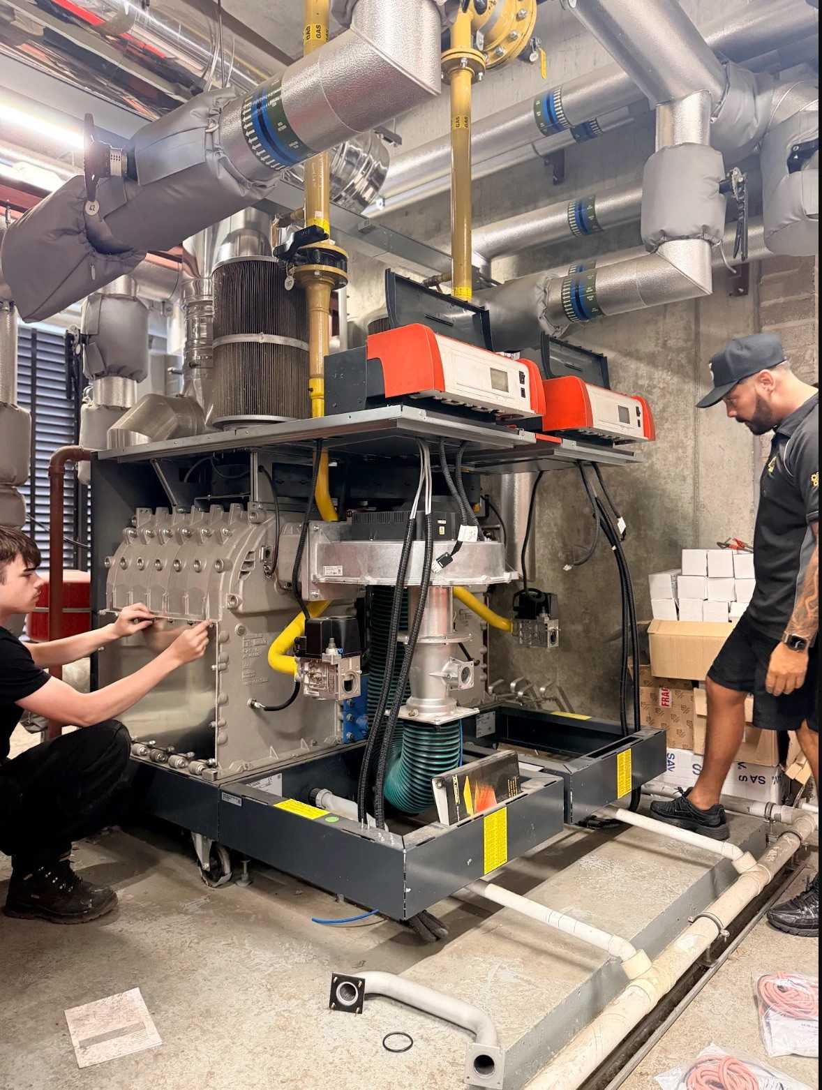 Two Neos Engineering engineers servicing commercial gas boiler plant in a London plant room, under insulated heating pipes and yellow gas pipework
