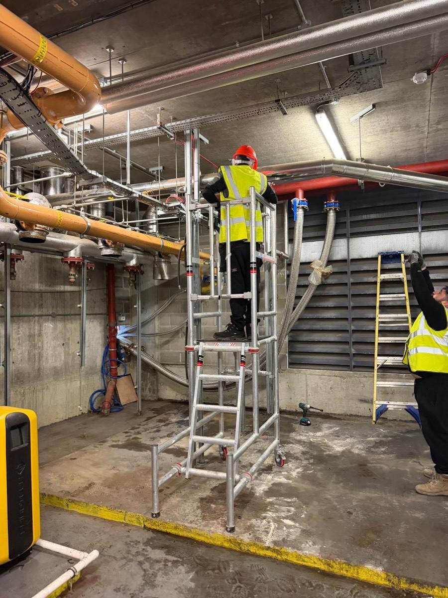 A Neos Engineering engineer on an access tower working on the pipework under a commercial plant room ceiling, with a second engineer at work beside it